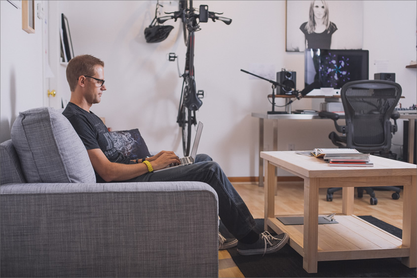 A photo of Cameron Moll sitting on the sofa in his office, using a laptop, with an iMac and speakers on the far right.